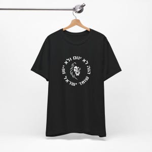 Op de afbeelding: Een zwart T-shirt met korte mouwen en een witte afbeelding op de borst. Het ontwerp toont een leeuwenkop in het midden van een cirkelvormige rand met Hebreeuwse tekst: הנה לא ינום ולא יישן שומר ישראל.