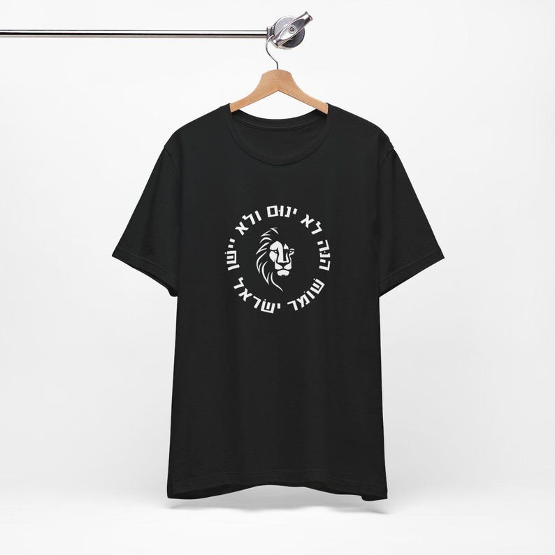 Op de afbeelding: Een zwart T-shirt met korte mouwen en een witte afbeelding op de borst. Het ontwerp toont een leeuwenkop in het midden van een cirkelvormige rand met Hebreeuwse tekst: הנה לא ינום ולא יישן שומר ישראל.