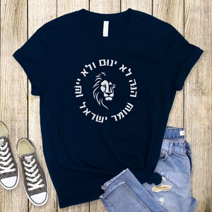 Op de afbeelding: Een marineblauw T-shirt met korte mouwen en een witte afbeelding van een leeuwenkop in het midden van een cirkel met Hebreeuwse tekst. De tekst "הנה לא ינום ולא יישן שומר ישראל" is in een vetgedrukt, schreefloos lettertype tegen de donkere stof.