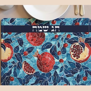 May include: A rectangular placemat featuring a mosaic-style design of pomegranates and leaves in shades of red, blue, and teal. The placemat has a dark blue band with Hebrew text. A white plate and silverware are partially visible.