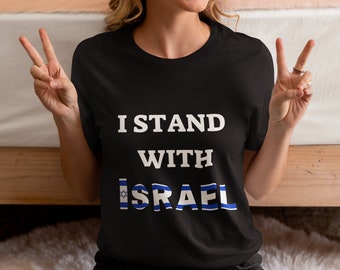 May include: Black t-shirt with the text "I STAND WITH ISRAEL" in white. The word "ISRAEL" is designed with the Israeli flag colors. The person is giving a peace sign with both hands.