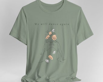 Op de afbeelding: Een saliegroen T-shirt met een lijntekening van een vrouwenhoofd met bloemen en vlinders eromheen. De tekst "We will dance again" staat op het T-shirt gedrukt.