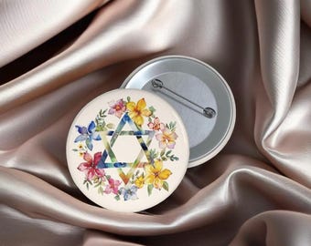 May include: A round button pin with a floral design and a Star of David in the center. The flowers are in shades of yellow, pink, blue, and purple. The pin is on a satin background.