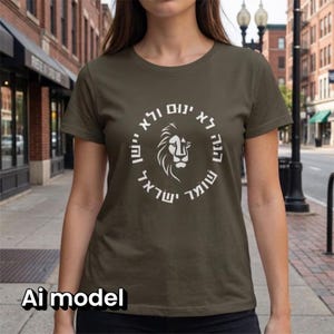 Op de afbeelding: Een olijfgroen T-shirt met korte mouwen en een witte cirkelvormige afbeelding op de borst. Het ontwerp bevat een gestileerde leeuwenkop in het midden, omringd door de Hebreeuwse tekst "הנה לא ינום ולא יישן שומר ישראל".