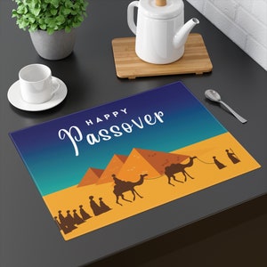 May include: A rectangular placemat with a desert scene featuring pyramids, camels, and figures. The words "Happy Passover" are written in white script against a gradient blue and yellow background. The placemat is on a dark surface.