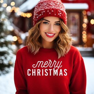 Op de afbeelding: Een vrouw draagt een rode trui met de tekst "merry christmas" in wit. Ze draagt een rode gebreide muts met een witte pompon.