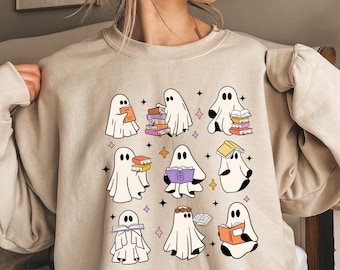 Puede incluir: Una sudadera beige con un estampado gráfico de fantasmas leyendo libros. Los fantasmas son blancos con contornos negros y sostienen libros de varios colores. El texto "Bookish Ghosts" está impreso en la sudadera.