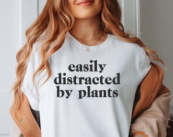 May include: White t-shirt with black text that reads "easily distracted by plants".