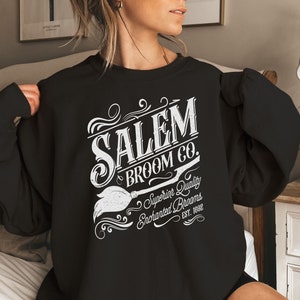 May include: Black sweatshirt with white text that reads "SALEM BROOM CO." with a broom graphic. The text also includes "Superior Quality Enchanted Brooms EST. 1692". The sweatshirt is a crew neck style.