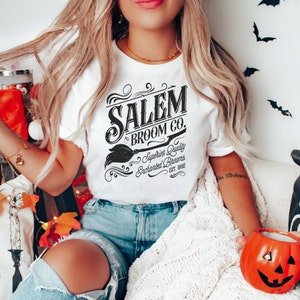 May include: White t-shirt with the text "Salem Broom Co." in a vintage black font, featuring a broom graphic. The shirt also includes the text "Superior Quality Enchanted Brooms Est. 1692". The model is wearing blue denim shorts.
