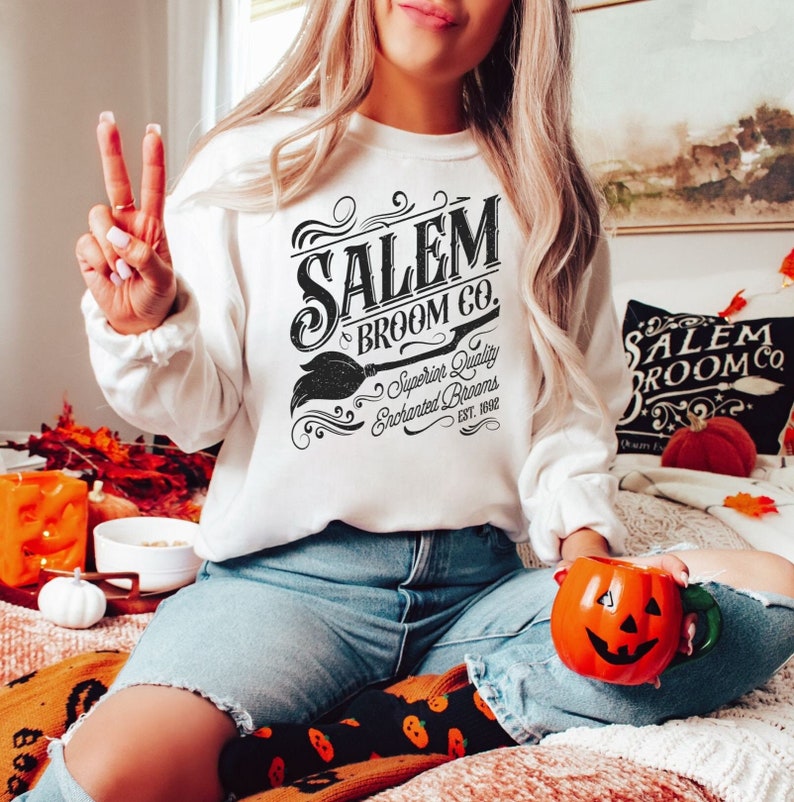 May include: A white sweatshirt with the text "Salem Broom Co." in black, featuring a broom graphic. The sweatshirt is part of a Halloween-themed collection. The person is wearing blue jeans and holding an orange pumpkin mug.