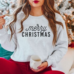 Op de afbeelding: Een grijs sweatshirt met de tekst "merry CHRISTMAS" in zwarte letters. Het sweatshirt wordt gedragen door een persoon die op een witte bank zit met een rood-wit geruite deken.