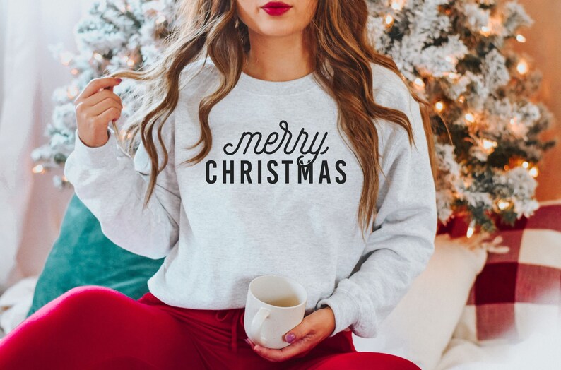 Op de afbeelding: Een grijs sweatshirt met de tekst "merry CHRISTMAS" in zwarte letters. Het sweatshirt wordt gedragen door een persoon die op een witte bank zit met een rood-wit geruite deken.