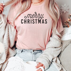 Op de afbeelding: Een vrouw draagt een roze T-shirt met de tekst "merry CHRISTMAS" in zwarte letters. Ze zit op een wit deken en houdt een witte mok vast.
