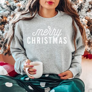 Op de afbeelding: Een grijs sweatshirt met de tekst "merry christmas" in witte letters. De persoon draagt een groene en witte stippen pyjama en houdt een witte mok vast.