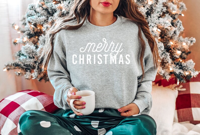 Op de afbeelding: Een grijs sweatshirt met de tekst "merry christmas" in witte letters. De persoon draagt een groene en witte stippen pyjama en houdt een witte mok vast.