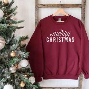 Op de afbeelding: Bordeauxkleurige trui met witte tekst die "merry christmas" zegt op een witte achtergrond. De trui hangt aan een houten ladder voor een kerstboom.