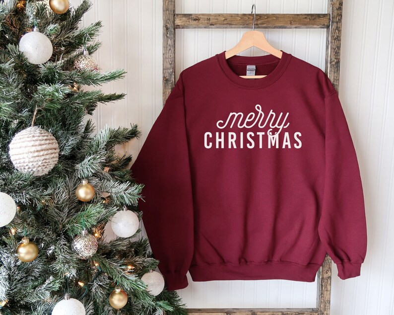 Op de afbeelding: Bordeauxkleurige trui met witte tekst die "merry christmas" zegt op een witte achtergrond. De trui hangt aan een houten ladder voor een kerstboom.