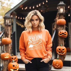 May include: An orange sweatshirt with the text "Salem Broom Co." in white, featuring a broom graphic. The sweatshirt is worn with black pants. The background includes carved pumpkins and a house decorated for Halloween.