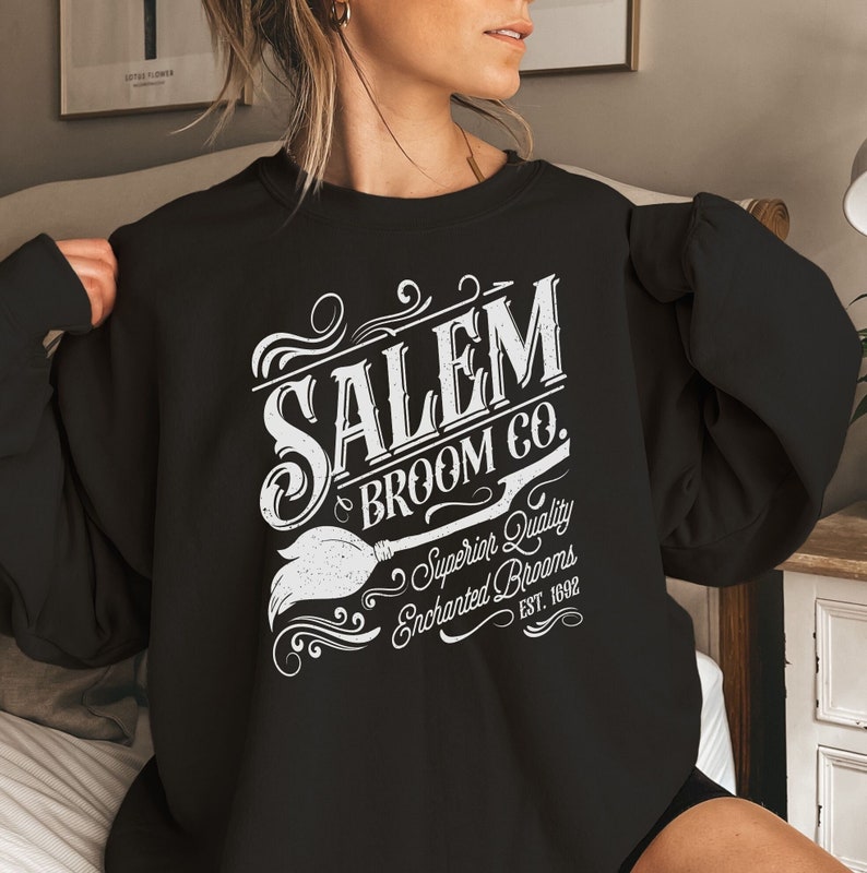 May include: Black sweatshirt with white text that reads "SALEM BROOM CO." with a broom graphic. The text also includes "Superior Quality Enchanted Brooms EST. 1692". The sweatshirt is a crew neck style.