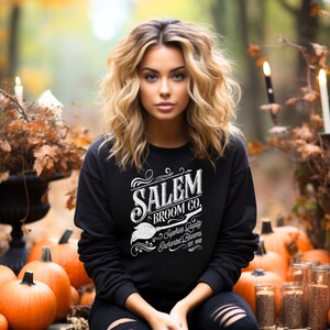 May include: A black sweatshirt with the text "SALEM BROOM CO." in white, featuring a broom graphic. The sweatshirt is worn by a person sitting among pumpkins and autumn foliage. The text also includes "Superior Quality Enchanted Brooms Est. 1692."