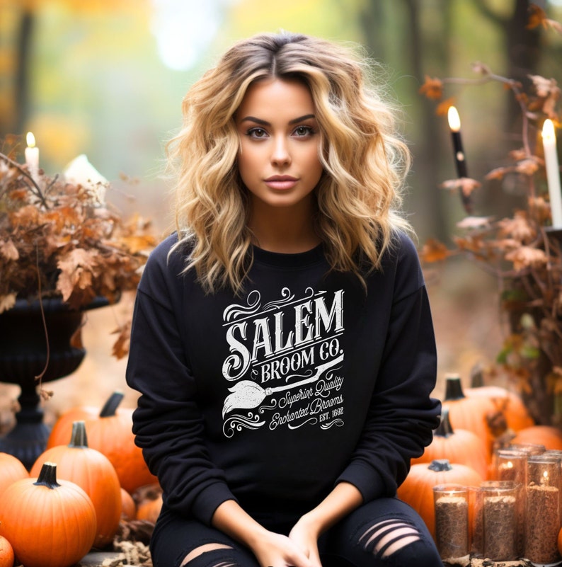 May include: A black sweatshirt with the text "SALEM BROOM CO." in white, featuring a broom graphic. The sweatshirt is worn by a person sitting among pumpkins and autumn foliage. The text also includes "Superior Quality Enchanted Brooms Est. 1692."
