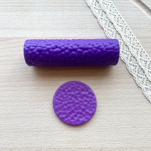 Può includere: Un rullo cilindrico viola e un disco circolare viola abbinato, entrambi caratterizzati da una superficie testurizzata e irregolare. Gli oggetti sono di colore solido e presentano un motivo in rilievo organico e ripetitivo su tutto l'esterno.