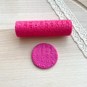 以下が含まれることがあります： 鮮やかなピンク色の円筒形ローラーと、お揃いの円形ディスク。どちらも古代エジプトのヒエログリフが深く刻印された模様が施されています。これらのツールは、粘土や生地などの柔らかい素材に質感のある型押しをするために設計されています。