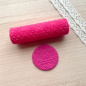 Può includere: Un mattarello rosa acceso strutturato e un timbro circolare abbinato. Il mattarello è cilindrico e presenta un motivo ripetuto. Un pezzo di pizzo bianco è sullo sfondo. Gli oggetti sono su una superficie di legno chiaro.