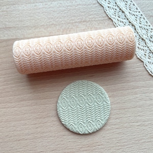May include: A beige rolling pin with a raised geometric pattern. The pattern is a repeating design of small circles and lines. The rolling pin is on a light brown wooden surface.