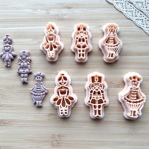 Può includere: Una collezione di formine per biscotti in plastica rosa di varie forme, tra cui una ballerina, un soldato e altre figure. Le formine sono disposte su una superficie di legno chiaro, con un bordo di pizzo decorativo sullo sfondo.
