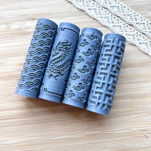May include: Four cylindrical grey rollers featuring distinct embossed patterns including repeating waves, a detailed dragon, stylised cloud motifs, and a geometric grid design. These textured rollers are designed for creating impressions in clay or soft materials.