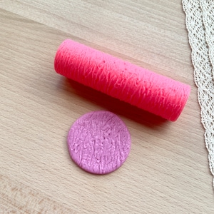 Puede incluir: Un rodillo de amasar rosa y un molde redondo de silicona morado con una superficie texturizada. Ambas herramientas se utilizan para hornear y decorar pasteles y otros postres.