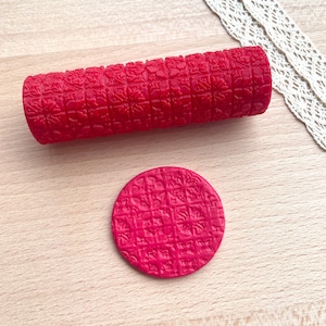 Peut inclure: Un rouleau à texture cylindrique rouge et un disque en argile rouge assorti. Les deux objets présentent un motif en grille répétitif avec des détails floraux complexes en relief sur la surface, créant une texture uniforme et précise.