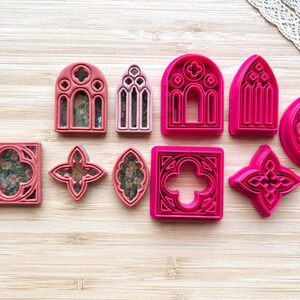 May include: A collection of ten plastic cookie cutters in various shapes including arched windows, squares, and quatrefoils. The cutters are colored in shades of pink and rose, with some containing floral patterned inserts within their frames.