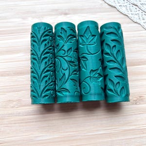 Könnte beinhalten: Vier teal-farbene zylindrische Walzen mit komplizierten Blumen- und Blättermustern. Die Walzen sind horizontal auf einer hellen Holzoberfläche angeordnet und zeigen detaillierte botanische Designs. Eine weiße Spitzentrimmung ist im Hintergrund sichtbar.