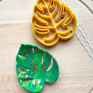 May include: Two monstera leaf shaped cookie cutters. One is yellow plastic and the other is green and gold painted clay.