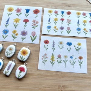 Könnte beinhalten: Drei rechteckige Blätter aus weißem Papier mit verschiedenen floralen Illustrationen im Aquarellstil sowie sechs einzelne Anhänger mit Metallrand, die jeweils ein einzelnes gemaltes Blumenmotiv in Blau, Gelb, Rot und Violett zeigen.