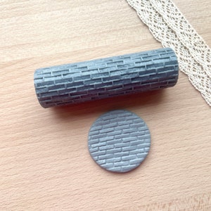 May include: A cylindrical gray rolling pin featuring a raised brick pattern texture, accompanied by a small, circular gray disc that displays the same embossed brick design on its surface, creating a consistent pattern for crafting applications.