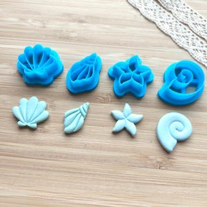 Può includere: Una collezione di formine per biscotti a forma di conchiglia blu e azzurre. Le formine includono vari disegni come una conchiglia, una stella marina e una conchiglia a spirale. Un nastro di pizzo decorativo è sullo sfondo.