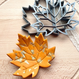May include: A grey plastic maple leaf cookie cutter and a matching orange maple leaf shape with gold leaf accents. The orange leaf features embossed vein details across its surface, while the grey cutter shows a sharp, defined outline.