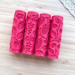 Può includere: Quattro rulli cilindrici rosa con motivi in rilievo di vari frutti e foglie. Ogni rullo presenta un design botanico distinto, tra cui fragole, uva, lamponi e mirtilli incisi sulla superficie del cilindro.