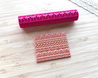 May include: A cylindrical pink rolling pin and a square pink stamp, both featuring an embossed pattern of repeating fan-like shapes and geometric Greek key borders. The stamp shows the raised design highlighted in a metallic gold color.