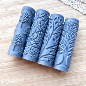 Op de afbeelding: Vier lichtblauwe, cilindrische kunststempels met boomontwerpen. Elke stempel heeft een ander boompatroon, inclusief gedetailleerde bladeren en takken. De stempels zijn gerangschikt op een licht houten oppervlak.