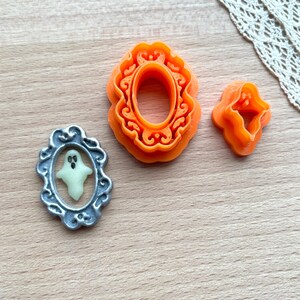 Op de afbeelding: Drie oranje plastic koekjesvormen in de vorm van sierlijke frames. Een frame heeft een spook erin.