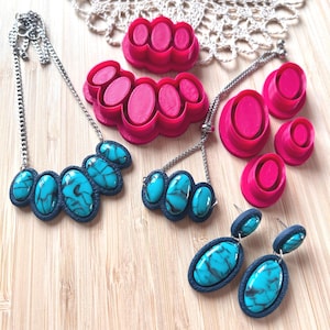 May include: A collection of jewellery featuring turquoise-coloured oval stones with black veining set in dark blue textured frames, including two necklaces and a pair of earrings, alongside several bright pink oval-shaped plastic cutters.