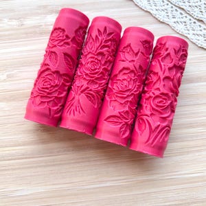 May include: Four vibrant pink, cylindrical embossing rollers with detailed rose and leaf patterns. The rollers are arranged diagonally on a light wood-grain surface, showcasing the floral design. A white lace trim is visible in the background.