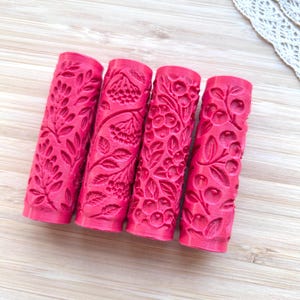 Op de afbeelding: Vier cilindervormige roze rollers met ingewikkelde botanische reliëfpatronen. Elke roller toont een uniek ontwerp van bladeren, bessen en bloemtrossen in het oppervlak, bedoeld voor het maken van texturen op zachte materialen.