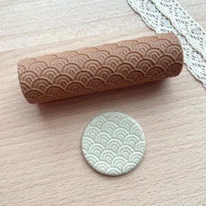 May include: A cylindrical wooden rolling pin featuring a deeply engraved repeating pattern of overlapping semicircles. Beside the rolling pin lies a small, circular, light-coloured clay disc that displays the same embossed scale-like texture.