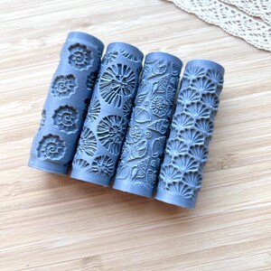 Könnte beinhalten: Vier blaue, strukturierte Nudelrollen mit verschiedenen Muschelmustern. Die Nudelrollen sind zylindrisch und weisen detaillierte Designs verschiedener Meereslebewesen auf, darunter Spiralmuscheln und fächerförmige Muster. Die Rollen sind diagonal auf einer hellen Holzoberfläche angeordnet.